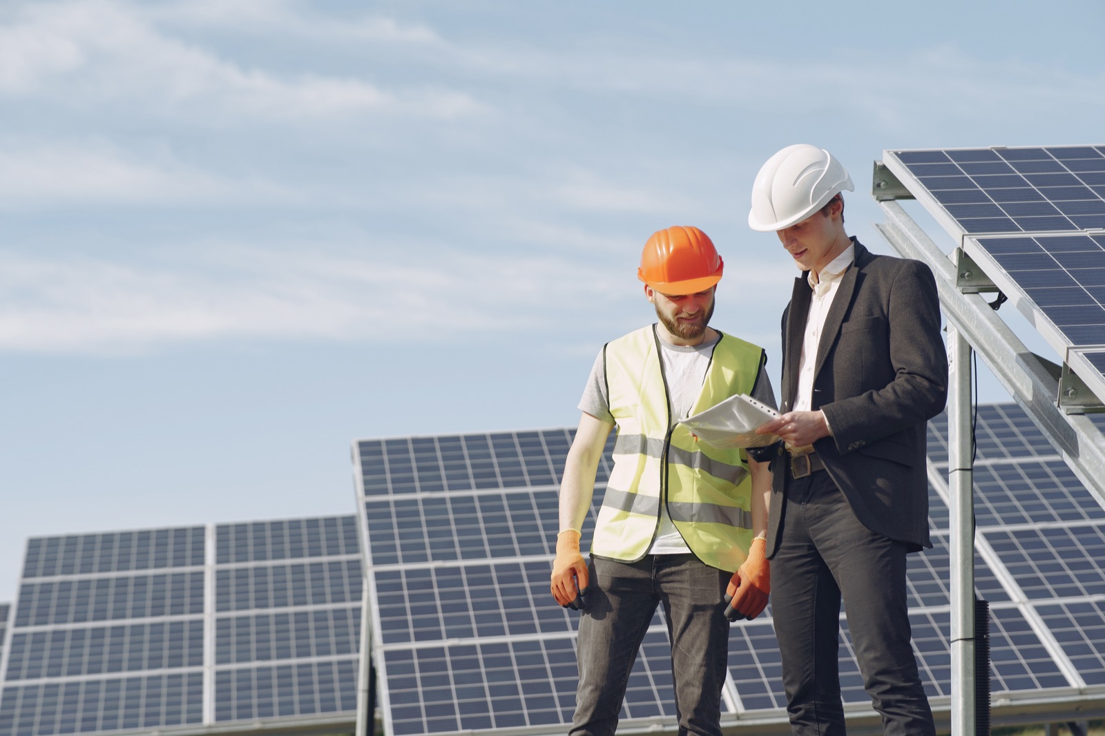 Two people wearing hard hats review documents beside solar panels